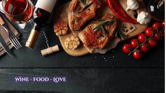 A bottle of red wine next to a glass of red wine on a table with grilled meat, garlic cloves, cherry tomatoes and cutlery